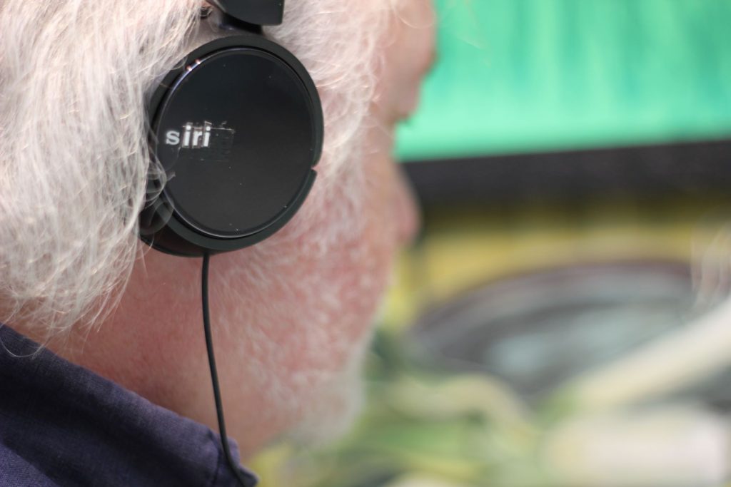 Close up and side on view of Artist Chris Tally Evans, wearing a set of black headphones that are marked with the word 'Siri', looking at the exhibition, 'Thinking Green' by Owen Griffiths 