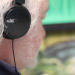Close up and side on view of Artist Chris Tally Evans, wearing a set of black headphones that are marked with the word 'Siri', looking at the exhibition, 'Thinking Green' by Owen Griffiths