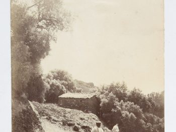 Sepia photo from North Africa Travel Album of two people in the foreground walking away from the camera, on a sloped dusty track. In front of them is a small stone single story building, surrounded by bushes and trees to the left.