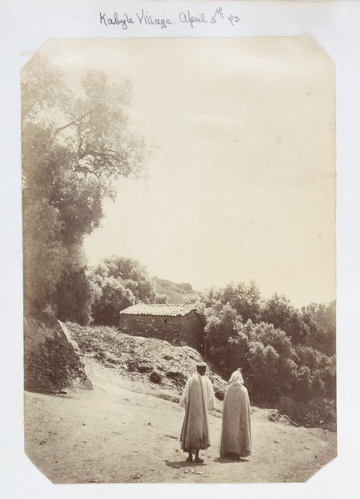 Sepia photo from North Africa Travel Album of two people in the foreground walking away from the camera, on a sloped dusty track. In front of them is a small stone single story building, surrounded by bushes and trees to the left.