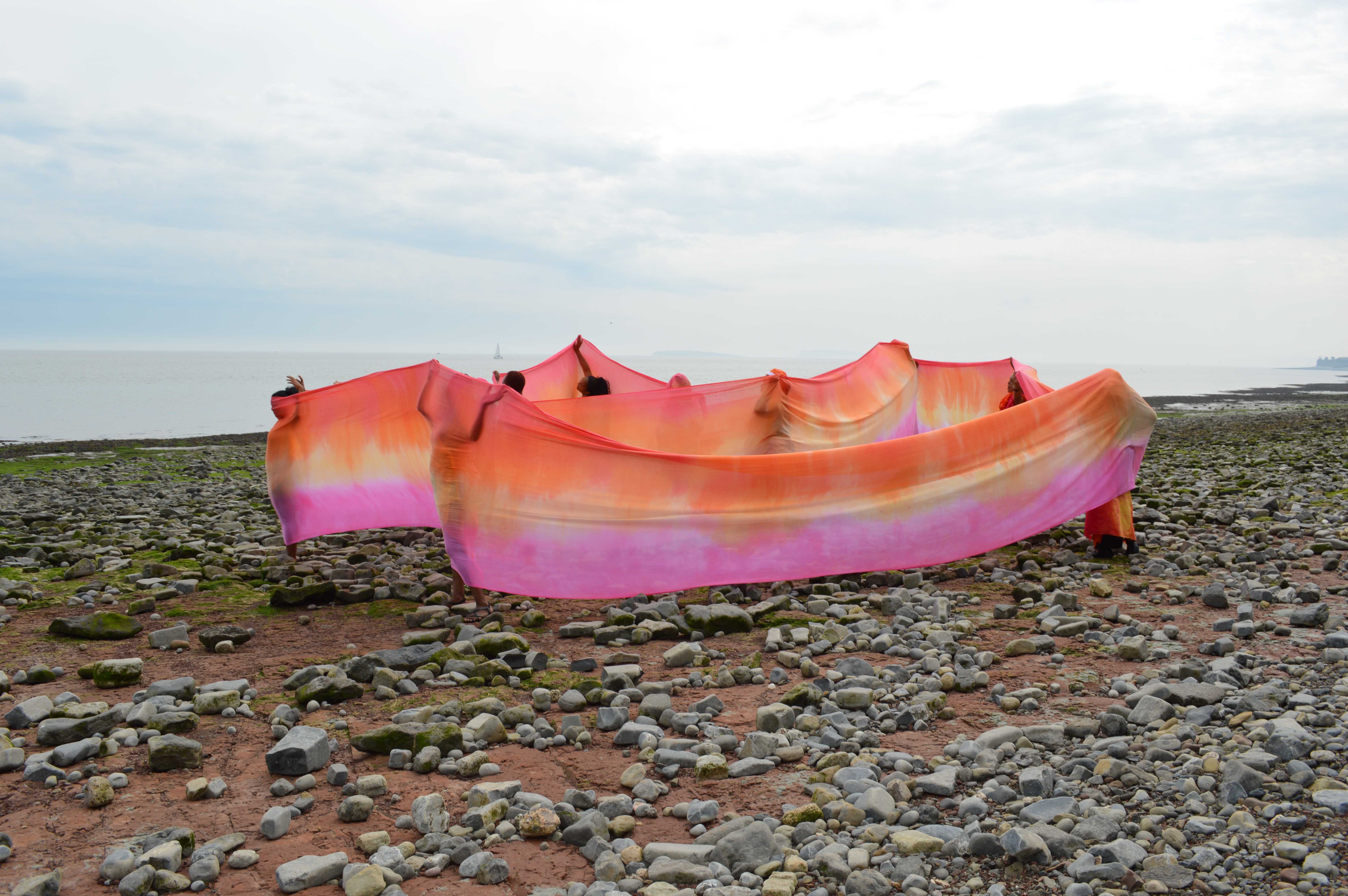 Women holding a large piece of dyed orange and pink fabric, wrapped around their bodies. Standing on a pebbled beach, grey skies in the background.