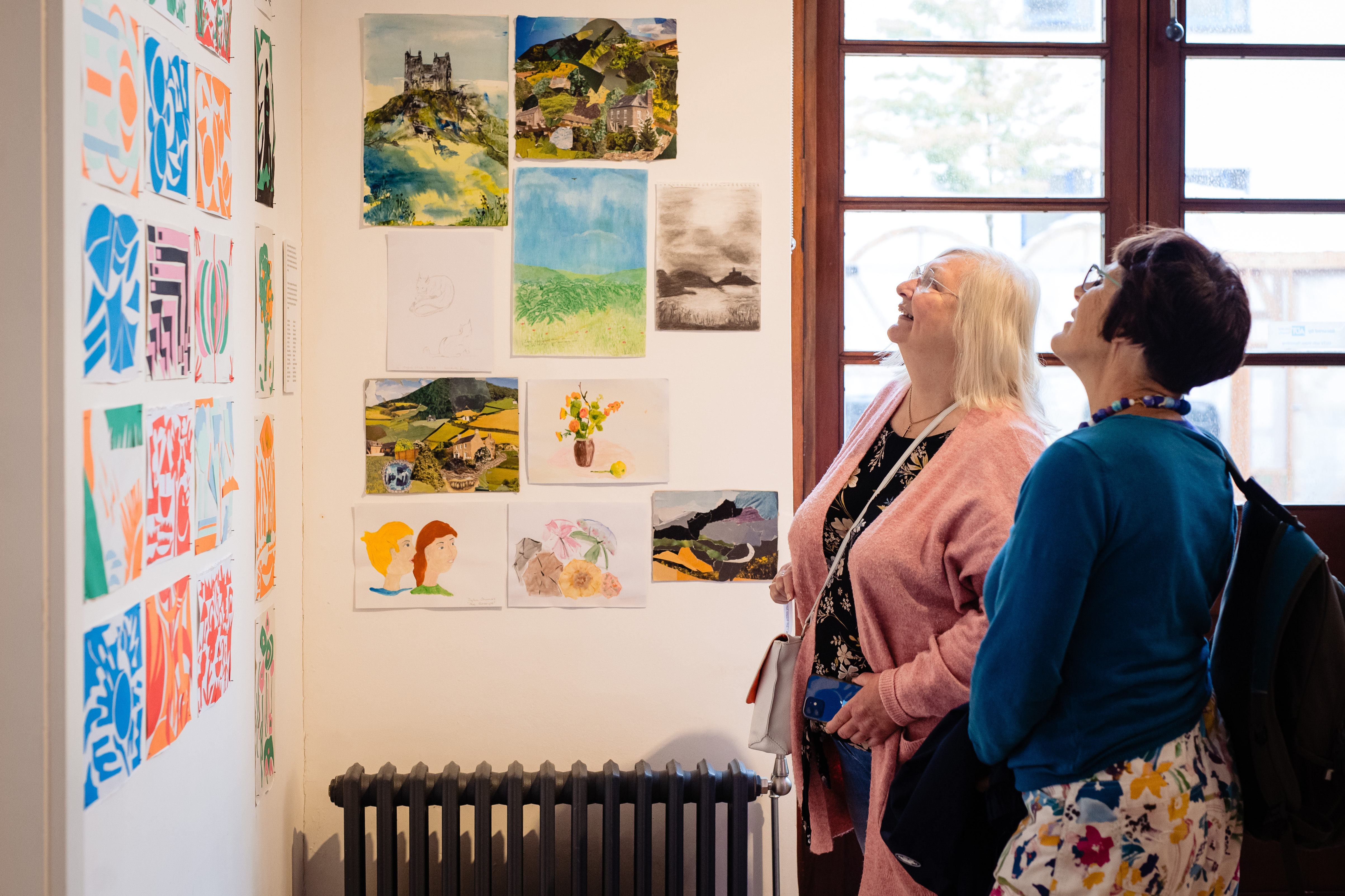 Side view of two ladies, looking up at a display of artwork on the left hand wall