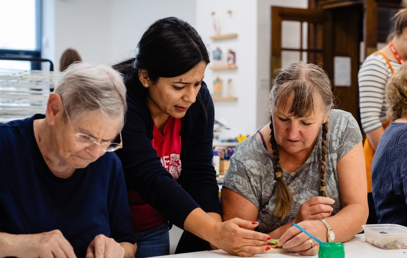 Three women looking down at a table. The women in the middle is demonstrating something to the woman on right. They are taking part in an art workshop