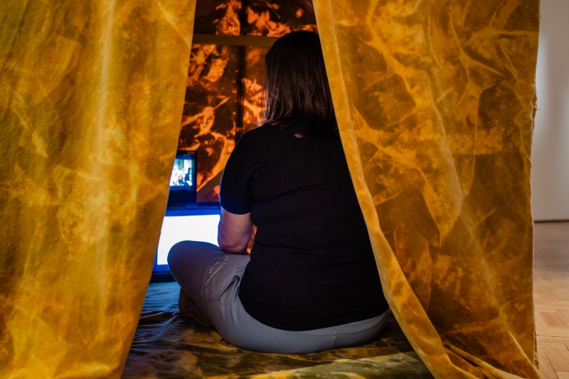 The back of someone sitting down inside a orange tent. The person inside the tent is watching two old style screens