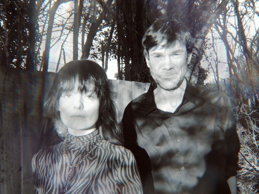 A black and white photograph of a man and woman looking directly to camera. They are outside, and there are trees in the background.