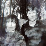 A black adn white photogrpah of a man and woman looking directly to camera. They are outside and there are trees in the background.